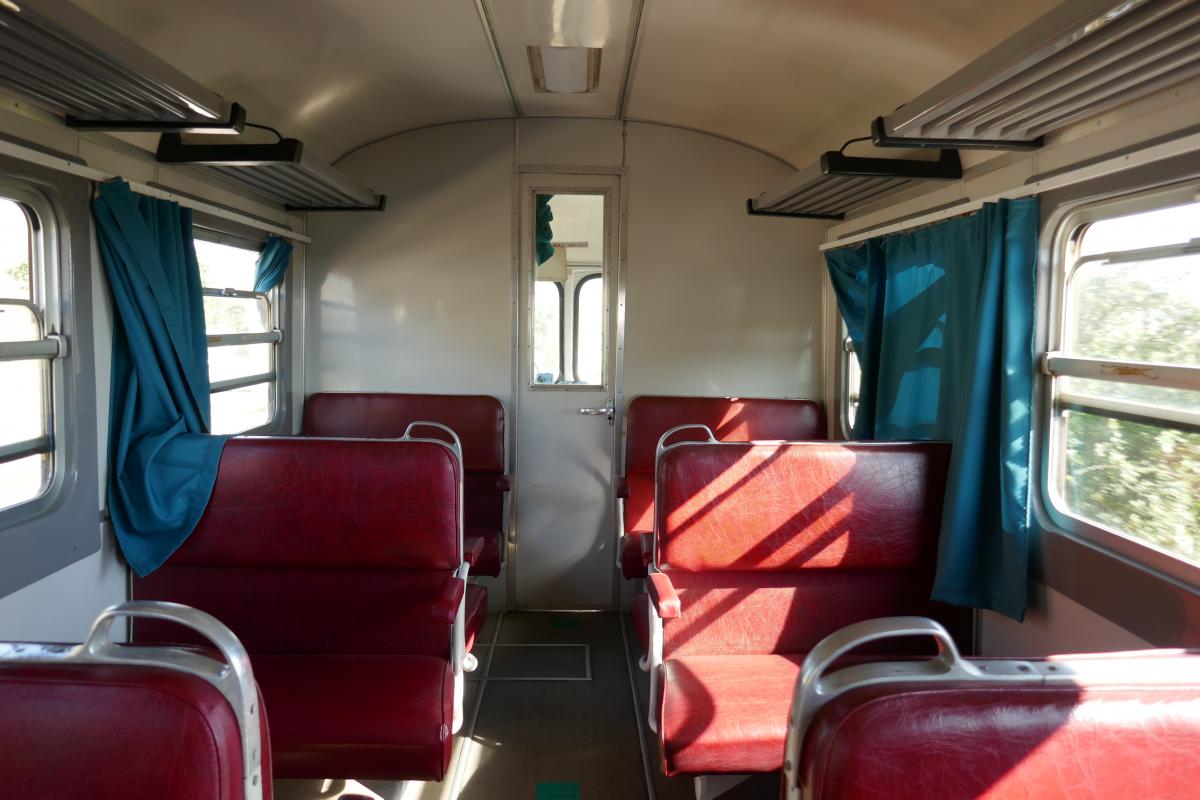 Interior of an old train carriage, empty, inundated by sun, open windows with blue curtains flowing in the wind, red vinil-covered seats