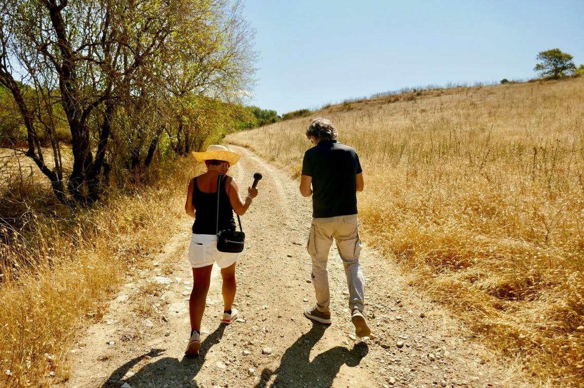 Cristina and Stefano are walking ahead, we see them from the back while they are talking, Crisitna is holding a microphone interviewing Stefano, they are walking in a sunny, dry landscape