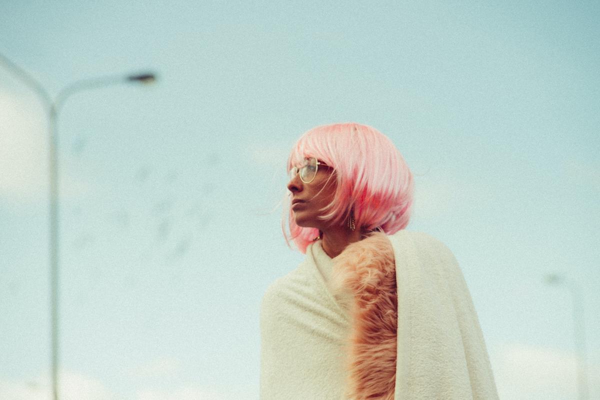 woman with red wid, short bob, wearing metallic glasses, looking into the distance in front of soft focus blue sky