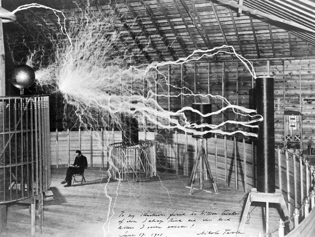 Nikola Tesla seated in a lab, surrounded by electrical equipment as bolts of lightning arc from a Tesla coil. Date: June 17, 1891.